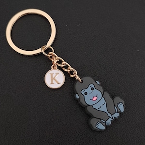 May include: Gold-tone keychain featuring a cartoon gorilla charm with a pink tongue and a white and gold initial charm. The keychain has a gold ring and chain.