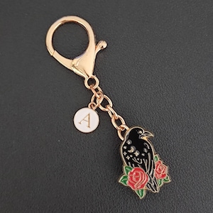 May include: Gold-coloured keychain featuring a black raven design, red roses, and a white initial charm. The raven has a crescent moon and stars design. The keychain has a lobster clasp.