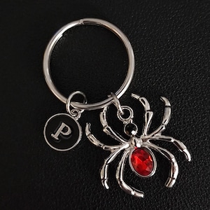 May include: Silver-coloured keyring featuring a spider charm with a red gem centre and a circular charm with the letter "P". The keyring includes a large ring for keys.