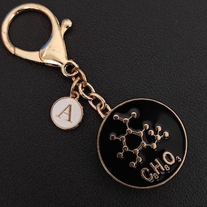 Op de afbeelding: Goudkleurige sleutelhanger met een zwarte, ronde bedel met een moleculaire structuur en de chemische formule C8H8O3. Inclusief een kleinere witte bedel met de letter "A". De sleutelhanger heeft een karabijnhaaksluiting.