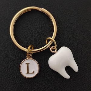 May include: Gold-coloured keyring featuring a white tooth charm and a round charm with the letter "L". The keyring is set against a black backdrop. The tooth charm is a detailed molar.