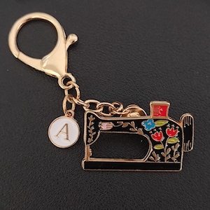 May include: A gold-coloured keyring featuring a sewing machine charm. The black sewing machine is decorated with colourful floral designs and has a gold-coloured outline. A white circular charm with the letter "A" is also attached.