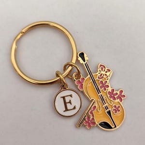 Puede incluir: Llavero dorado con un dije de violín amarillo decorado con flores rosas y un dije de círculo blanco con la letra E.