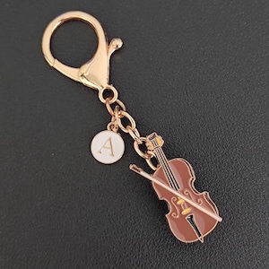 May include: A gold-colored keychain with a violin charm and a round charm with the letter "A". The violin is brown with gold accents and a bow. The keychain has a lobster clasp.