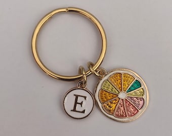 May include: Gold-tone keychain with a round charm and a citrus slice charm. The round charm has the letter "E" in white. The citrus slice charm features a colorful design with shades of yellow, green, pink, and orange.