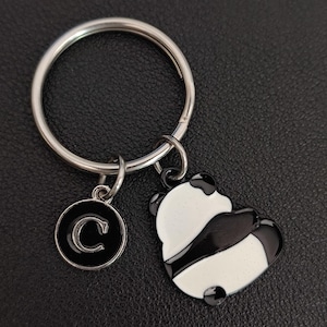 May include: Silver keyring with a panda bear charm and a circular charm with the letter "C". The panda charm is black and white. The keyring is on a black surface.