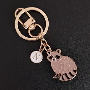 May include: A gold-coloured keyring featuring a raccoon charm with black and brown details. The keyring includes a round charm with the letter "N" in white. The background is black.