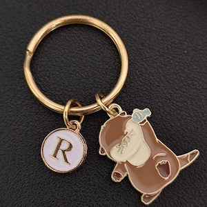 May include: A gold-coloured keyring with a round charm featuring the letter "R" in black on a white background. The keyring also includes a brown and cream-coloured otter charm holding a small blue fish.