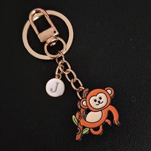 May include: A gold-coloured keyring featuring a cartoon monkey charm. The monkey is orange and tan, clinging to a brown branch with green leaves. A small white circular charm with the letter "J" hangs nearby.