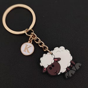 May include: A gold-coloured keyring featuring a cartoon sheep charm. The sheep has a white fluffy body, a brown face and legs, and a pink inner ear. A small round charm with the letter "K" is also attached.