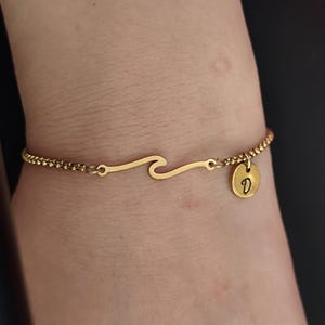 May include: A gold-toned bracelet featuring a central wave-shaped charm and a small circular pendant engraved with the letter D. The bracelet is constructed with a delicate, textured chain that connects to both sides of the wave design.
