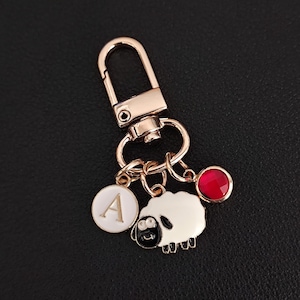 May include: A gold-tone keyring with a lobster clasp. It features a white sheep charm with a black face, a round charm with the letter "A", and a red gem. The background is black.