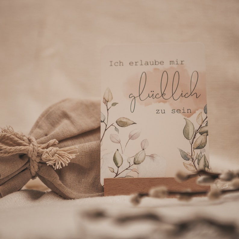May include: A white card with a watercolor floral design and the text "Ich erlaube mir gl&uuml;cklich zu sein" in black script. The card is propped up on a wooden stand and sits on a light brown surface.