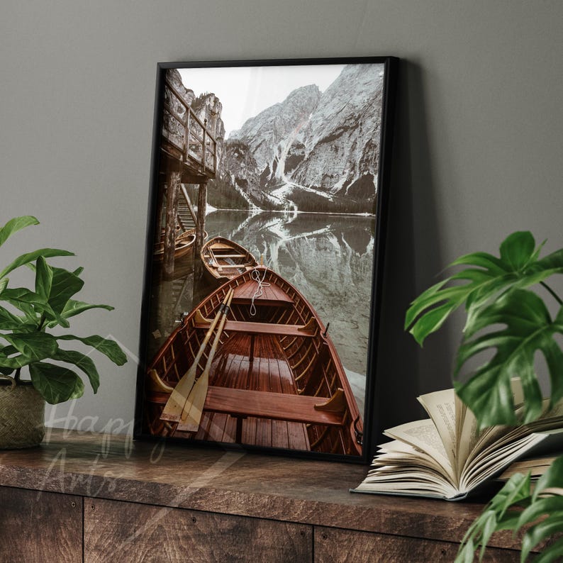 May include: A framed photograph of a lake with snow-capped mountains in the background. Two wooden rowboats are docked at a wooden pier. The boats are facing the camera, and the oars are visible.