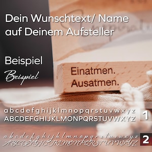 May include: A wooden block with the text "Einatmen. Ausatmen." engraved on it. The block is sitting on a beige fabric surface. The image also shows a font chart with lowercase and uppercase letters in two different styles.