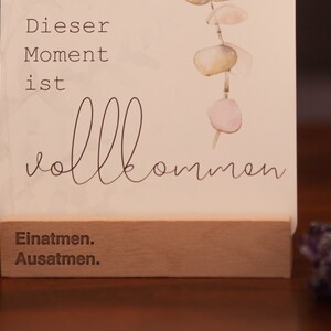 May include: A white card with the text "Dieser Moment ist vollkommen" and "Einatmen. Ausatmen." printed on it. The card is standing on a wooden base and has a watercolor illustration of a eucalyptus branch with pink and green leaves.