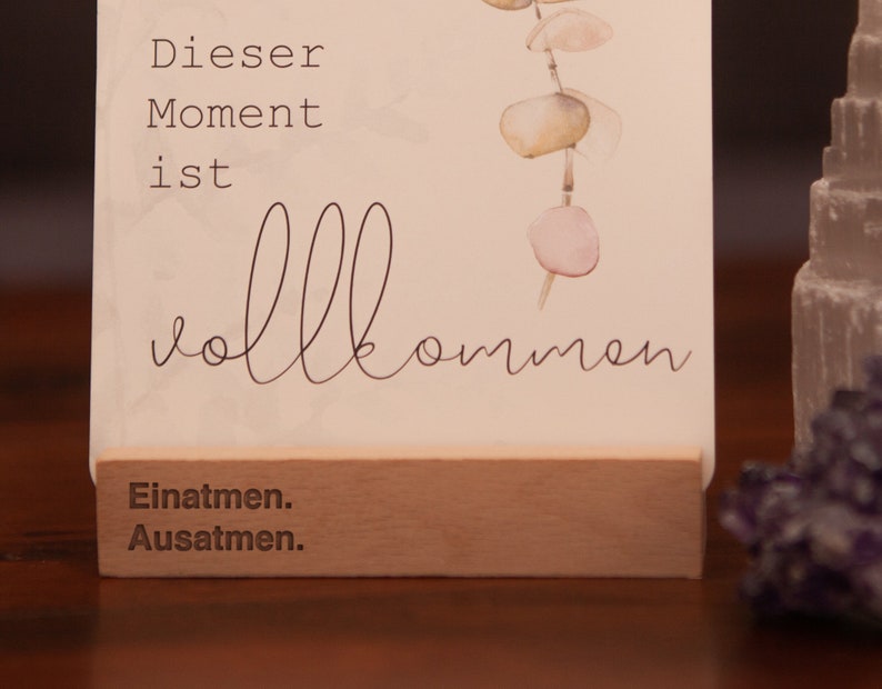 May include: A white card with the text "Dieser Moment ist vollkommen" and "Einatmen. Ausatmen." printed on it. The card is standing on a wooden base and has a watercolor illustration of a eucalyptus branch with pink and green leaves.