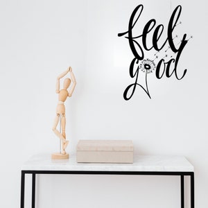 Op de afbeelding: Een houten, gearticuleerde pop die op een tafel staat met een witte doos en een zwart-witte muursticker met de tekst "feel good" en een paardenbloem afbeelding.