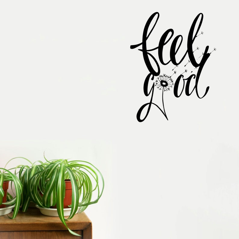 Op de afbeelding: Zwart-witte wanddecoratie met de tekst "feel good" in een cursief schrift. Een paardenbloem met zaden die in de wind waaien is in het ontwerp opgenomen.