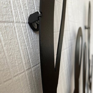 Op de afbeelding: Close-up van een zwarte metalen wanddecoratie met een decoratief ontwerp. De decoratie is bevestigd aan een witte muur met een zwarte clip.