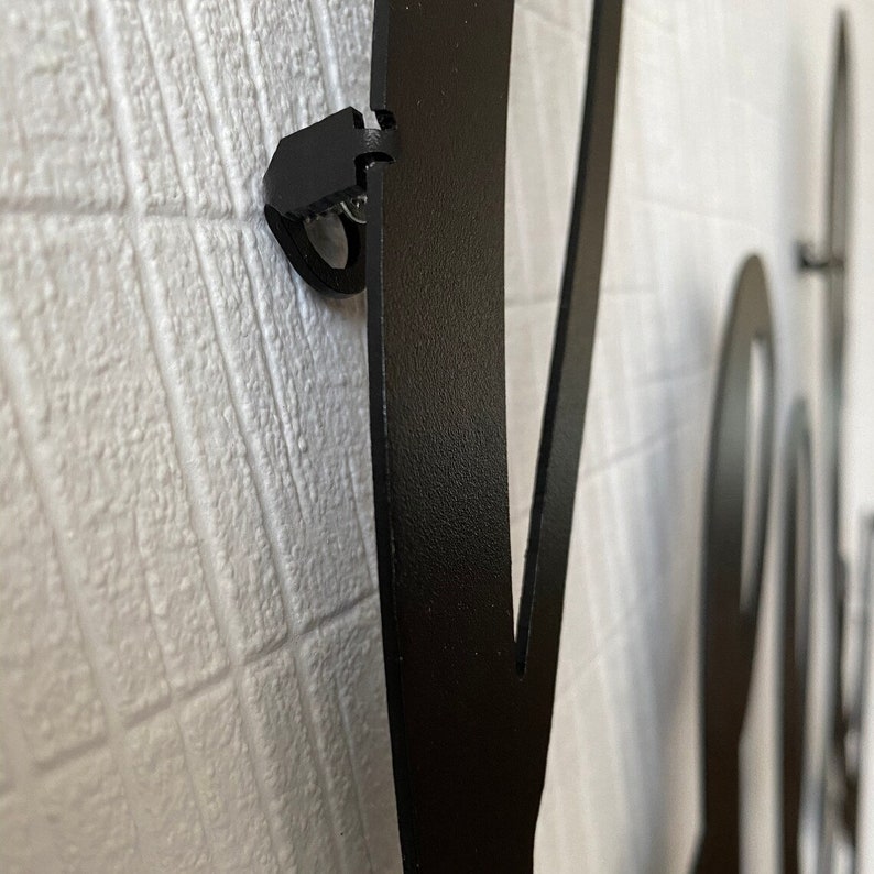 Op de afbeelding: Close-up van een zwarte metalen wanddecoratie met een decoratief ontwerp. De decoratie is bevestigd aan een witte muur met een zwarte clip.