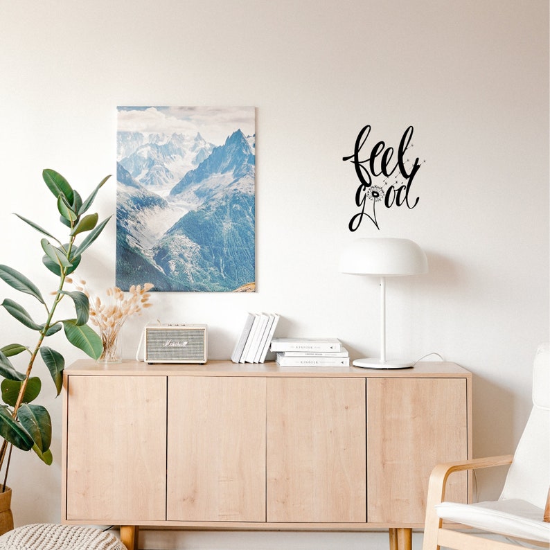 Op de afbeelding: Een licht houten kast met drie deuren en een witte lamp erop. Een ingelijst afdruk van een bergketen hangt aan de wand boven de kast. De tekst "feel good" is in zwarte cursieve letters op de wand geschreven met een paardenbloemontwerp.