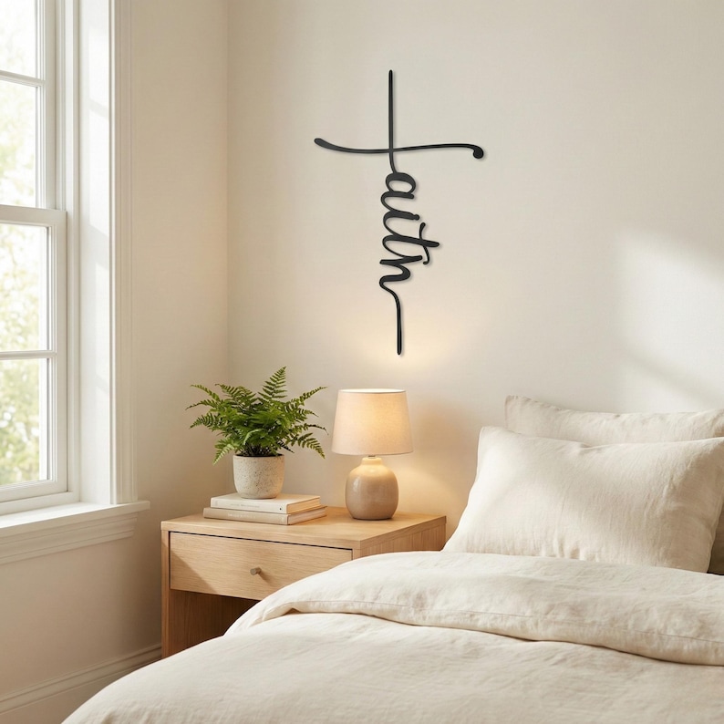 a serene bedroom scene with a bed, nightstand, and a potted plant. The bed is covered in a white comforter and has a single pillow. The nightstand has a lamp and a stack of books on it. Above the nightstand, there is a black metal cross-shaped sign with the word "Faith" written on it.