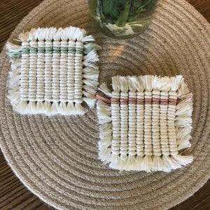 May include: Two handmade macrame coasters with fringe. One coaster has a green stripe and the other has a pink stripe.