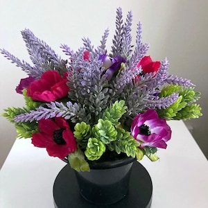 May include: A floral arrangement in a black pot features a mix of artificial flowers. The arrangement includes deep red poppies, purple anemones, and lavender sprigs, with green foliage accents. The pot sits on a black base.