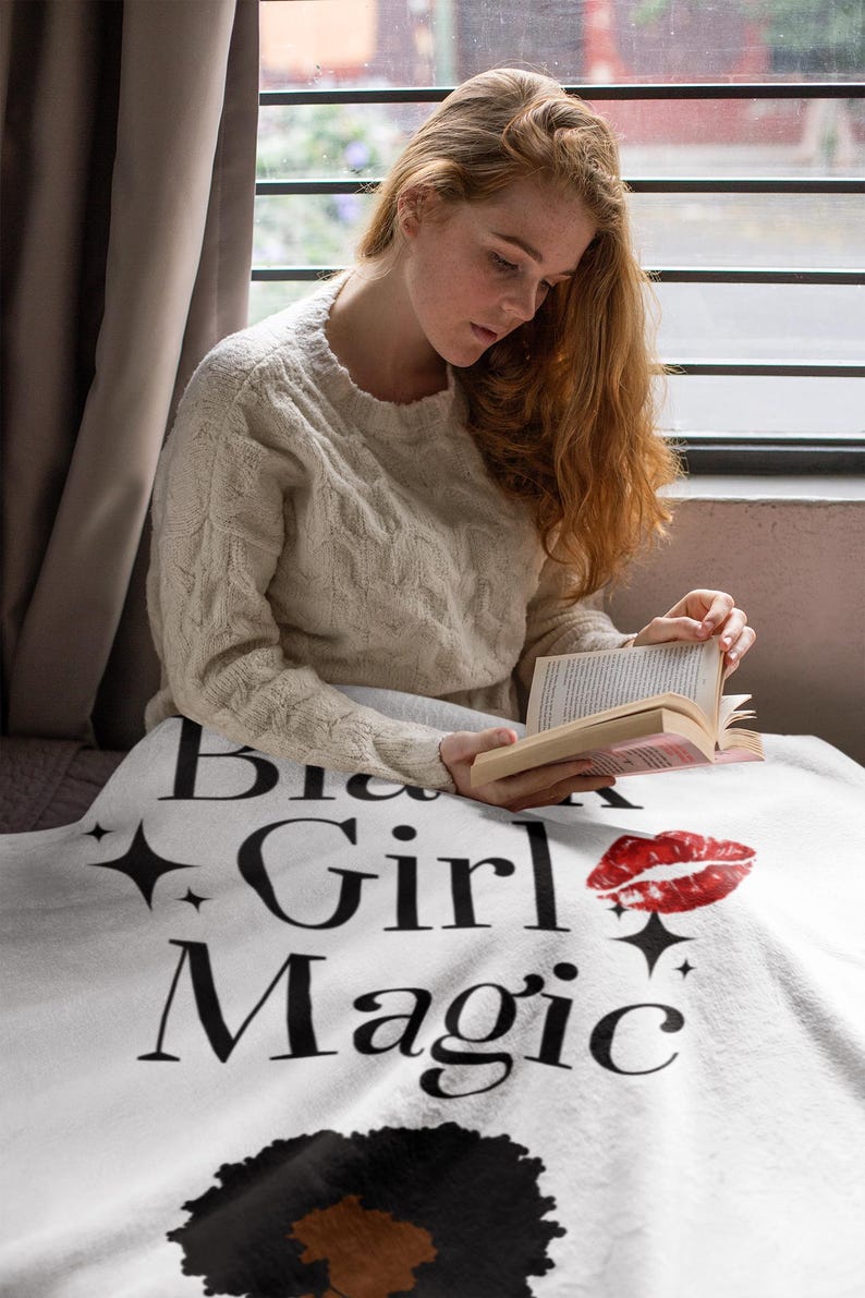 May include: A white blanket with the words "Big Girl Magic" in black script, a red lipstick kiss, and a black afro graphic. The blanket is draped over a person reading a book.