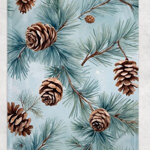 May include: A light blue blanket with a watercolor design of pine branches and brown pine cones. The blanket features a repeating pattern of the natural elements, creating a cozy and seasonal aesthetic.