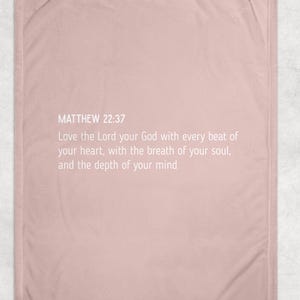 May include: A pale pink blanket featuring the text "MATTHEW 22:37" and a religious quote. The blanket has a plain design and a soft texture. The text is in a simple, easy-to-read font.