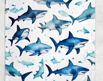 May include: A white blanket featuring a pattern of various blue sharks. The sharks vary in shades of blue, with some appearing darker and others lighter, creating a watercolor effect. The design includes both large and small sharks, along with smaller fish.