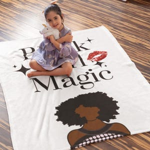 May include: A white blanket with the words "Pink and Magic" in black, a red lip print, and a black illustration of a woman's silhouette. A young girl is sitting on the blanket, holding a white unicorn plush toy.