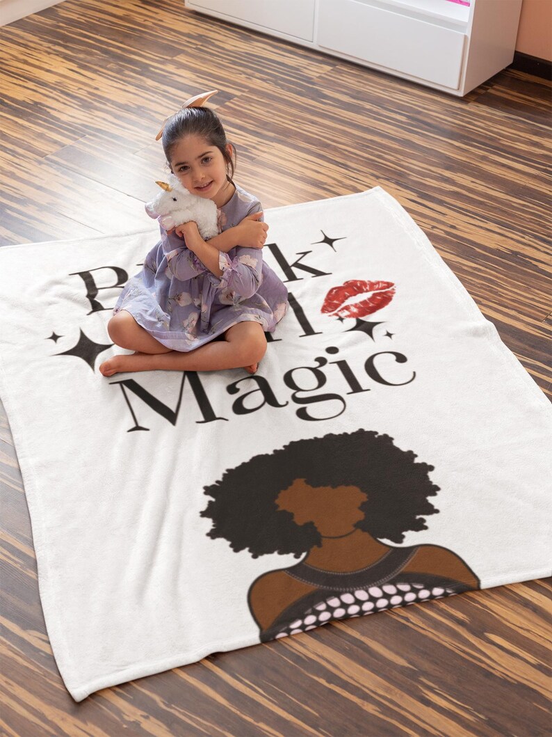 May include: A white blanket with the words "Pink and Magic" in black, a red lip print, and a black illustration of a woman's silhouette. A young girl is sitting on the blanket, holding a white unicorn plush toy.