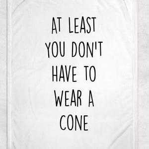 May include: White throw blanket with the text "AT LEAST YOU DON'T HAVE TO WEAR A CONE" in black, centered on the blanket. The blanket has a soft, textured appearance, suitable for home decor.