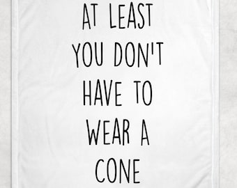 May include: White throw blanket with the text "AT LEAST YOU DON'T HAVE TO WEAR A CONE" in black, centered on the blanket. The blanket has a soft, textured appearance, suitable for home decor.