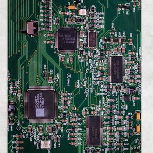 May include: Close-up of a green circuit board with intricate electronic components. The board features various chips, resistors, and connectors, with text labels and codes visible. The overall design is a complex network of pathways and solder points.