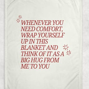 May include: Cream-coloured blanket with red text that reads "WHENEVER YOU NEED COMFORT, WRAP YOURSELF UP IN THIS BLANKET AND THINK OF IT AS A BIG HUG FROM ME TO YOU." Decorative starbursts accent the text.