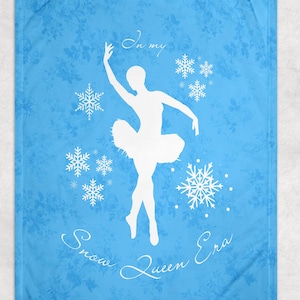 May include: A light blue blanket featuring a white silhouette of a ballerina, surrounded by snowflakes. The text "In my Snow Queen Era" is written in a cursive font. The blanket has a floral pattern.