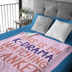 May include: Pink fleece blanket with the text "This is my K-Drama watching blanket" in pink, orange, and purple. The blanket has a small pink heart hand gesture in the corner.