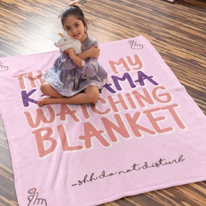 May include: Pink fleece blanket with the text "This is my K-drama watching blanket" in purple and orange lettering. The text "...Shh do not disturb" is printed in black at the bottom. A small girl is sitting on the blanket holding a stuffed unicorn.
