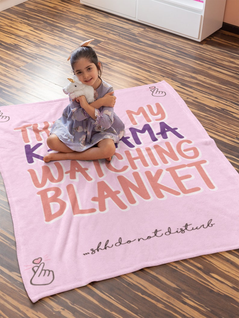 May include: Pink fleece blanket with the text "This is my K-drama watching blanket" in purple and orange lettering. The text "...Shh do not disturb" is printed in black at the bottom. A small girl is sitting on the blanket holding a stuffed unicorn.