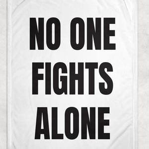May include: White blanket with the words "NO ONE FIGHTS ALONE" in bold, black capital letters. The text is centered and takes up most of the blanket's surface. The blanket has a soft, textured appearance.