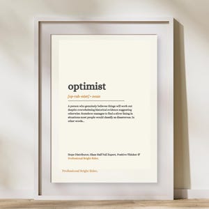 May include: A framed wall art print featuring a dictionary-style definition of the word optimist. The text includes the pronunciation, part of speech, a descriptive paragraph, and the phrases Hope Distributor, Glass Half Full Expert, and Professional Bright Sider in black and orange text on a cream background.