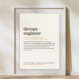 May include: A framed wall print featuring a dictionary-style definition of a devops engineer. The text is printed in dark gray and orange on a cream background, describing the role as responsible for automating deployments and managing infrastructure.