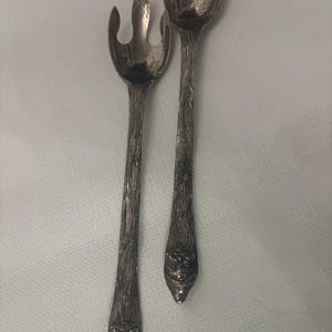 May include: A silver-toned fork and spoon set. The handles have a textured, tree-like design, and the ends feature a face. The fork has three tines. The spoon has a rounded bowl. The set is likely vintage or antique.