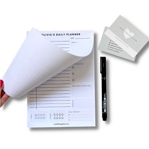 May include: A white daily planner with the text "OLIVIA'S DAILY PLANNER" at the top. The planner has sections for notes, priorities, new dental knowledge, exercise, and a meal tracker. A black marker pen and two business cards are also shown.