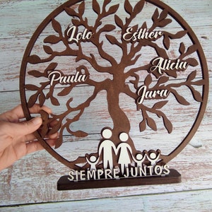 以下が含まれることがあります： 白い切り抜き葉と名前がついた茶色の木製のファミリーツリー。名前は「Lola」、「Esther」、「Alicia」、「Paula」、「Jara」です。木のベースには、白い切り抜きで家族が描かれており、「Siempre Juntos」という文字が書かれています。