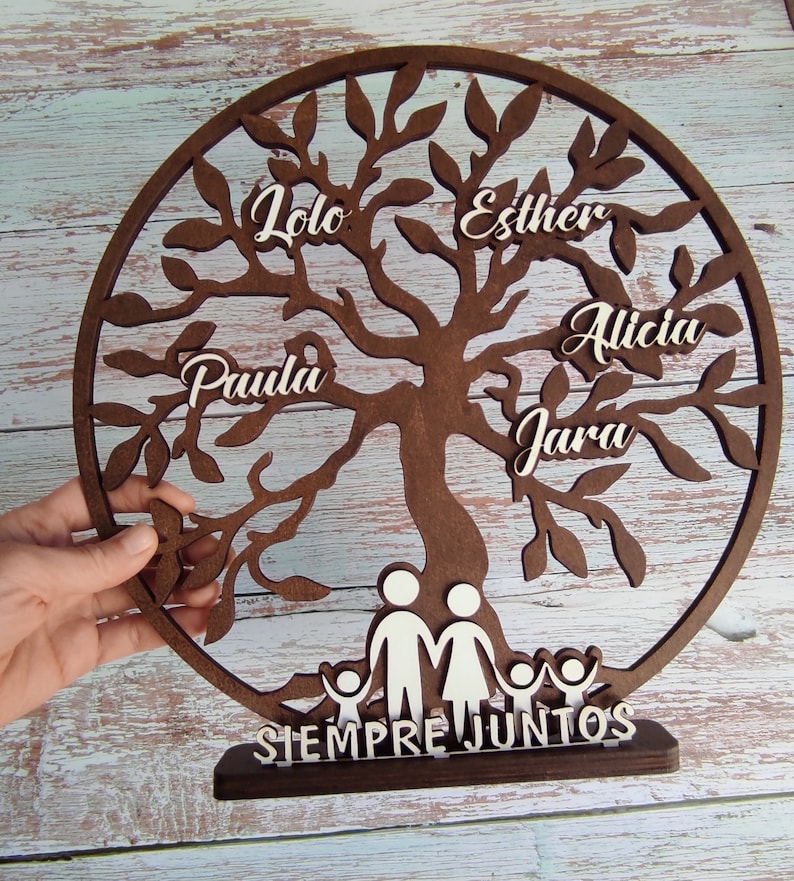以下が含まれることがあります： 白い切り抜き葉と名前がついた茶色の木製のファミリーツリー。名前は「Lola」、「Esther」、「Alicia」、「Paula」、「Jara」です。木のベースには、白い切り抜きで家族が描かれており、「Siempre Juntos」という文字が書かれています。